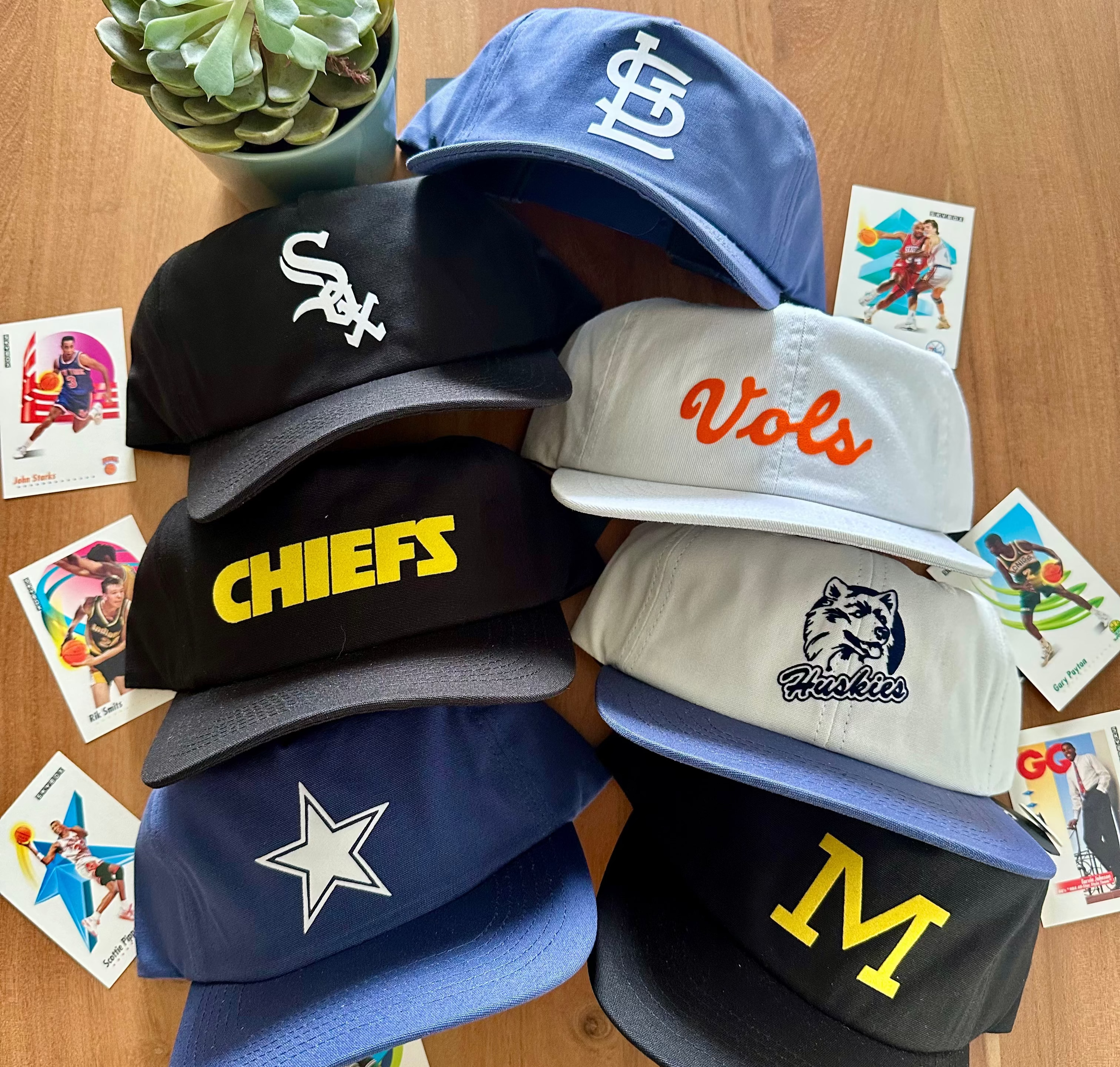 Collection of sports team hats on a wooden surface with small plants and cards.
