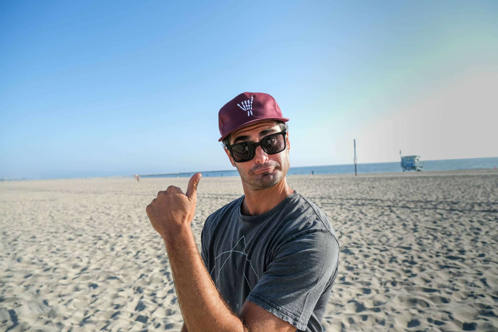 A man on a sandy beach wearing a maroon baseball cap with a white logo, black sunglasses, and a gray t-shirt, pointing toward the camera.